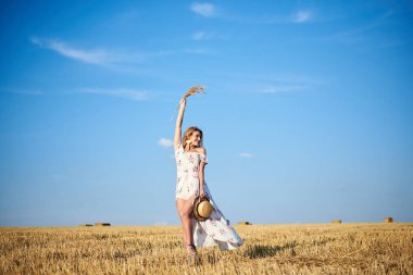 Genç sarışın kadın, beyaz romantik elbise ve hasır şapka giyiyor, elinde kuru çim buketi tutuyor, yazın saman tarlasında yürüyor. Doğal arka planda bir kadın portresi. Çevre koruma kavramı.