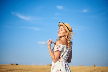 Genç sarışın kadın, beyaz romantik elbise ve hasır şapka giyiyor, elinde kuru çim buketi tutuyor, yazın saman tarlasında yürüyor. Doğal arka planda bir kadın portresi. Çevre koruma kavramı.