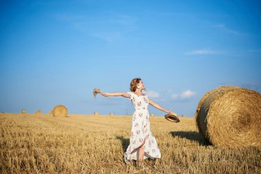 Beyaz romantik elbiseler giyen, elinde hasır şapka ve kurutulmuş çim buketi olan genç sarışın kadın yazın saman tarlasında zıplıyor. Doğal arka planda bir kadın portresi. Çevre koruma.