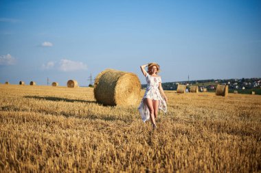Genç sarışın kadın, beyaz romantik elbise ve hasır şapka ve kurutulmuş çim buketi giyiyor, yazın saman tarlasında koşuyor. Doğal arka planda bir kadın portresi. Çevre koruma.