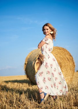 Sarışın, uzun, romantik bir elbise giyen, elinde hasır şapka ve kurutulmuş çimen buketi tutan, yazın tarlada buğday balyasının önünde poz veren genç bir kadın. Doğal arka planda kadın portresi.