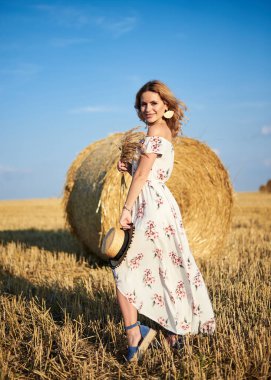 Sarışın, uzun, romantik bir elbise giyen, elinde hasır şapka ve kurutulmuş çimen buketi tutan, yazın tarlada buğday balyasının önünde poz veren genç bir kadın. Doğal arka planda kadın portresi.