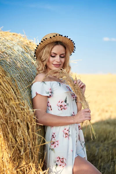 年轻的金发女人 穿着长长的白色浪漫连衣裙 头戴草帽 夏天靠在田里的麦兜上 自然背景的女性肖像 — 图库照片