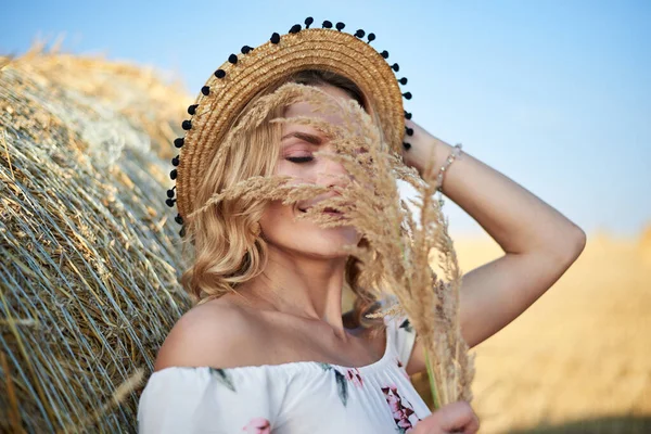 年轻的金发女人 穿着长长的白色浪漫连衣裙 头戴草帽 夏天靠在田里的麦兜上 自然背景的女性肖像 — 图库照片