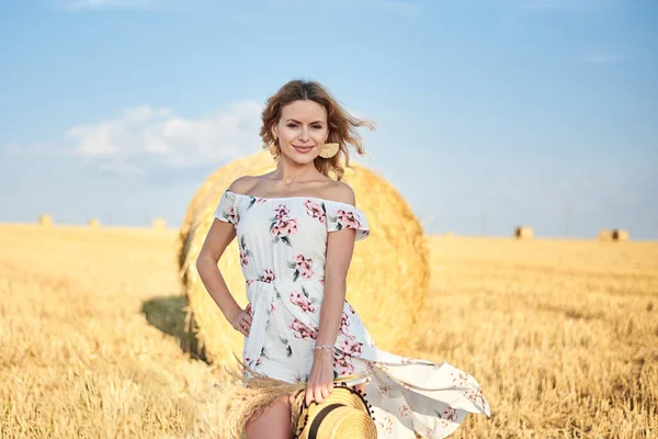 Sarışın, uzun, romantik bir elbise giyen, elinde hasır şapka ve kurutulmuş çimen buketi tutan, yazın tarlada buğday balyasının önünde poz veren genç bir kadın. Doğal arka planda kadın portresi.