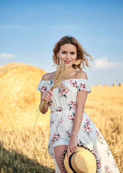 Sarışın, uzun, romantik bir elbise giyen, elinde hasır şapka ve kurutulmuş çimen buketi tutan, yazın tarlada buğday balyasının önünde poz veren genç bir kadın. Doğal arka planda kadın portresi.