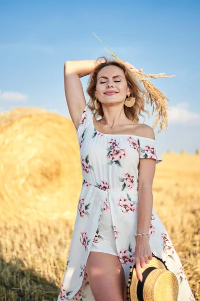 Sarışın, uzun, romantik bir elbise giyen, elinde hasır şapka ve kurutulmuş çimen buketi tutan, yazın tarlada buğday balyasının önünde poz veren genç bir kadın. Doğal arka planda kadın portresi.