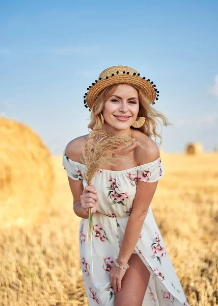 Sarışın, uzun, romantik bir elbise ve hasır şapka giyen, elinde kurutulmuş çim buketi tutan yazın tarlada buğday balyasının önünde poz veren genç bir kadın. Doğal arka planda kadın portresi.