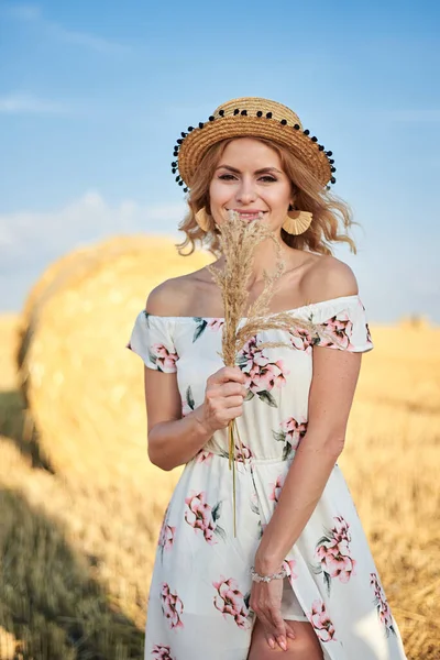 Sarışın, uzun, romantik bir elbise ve hasır şapka giyen, elinde kurutulmuş çim buketi tutan yazın tarlada buğday balyasının önünde poz veren genç bir kadın. Doğal arka planda kadın portresi.