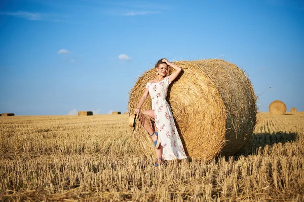 Genç sarışın kadın, uzun beyaz romantik elbise ve hasır şapka giyiyor, yazın tarlada buğday balyasına yaslanıyor. Doğal arka planda kadın portresi.
