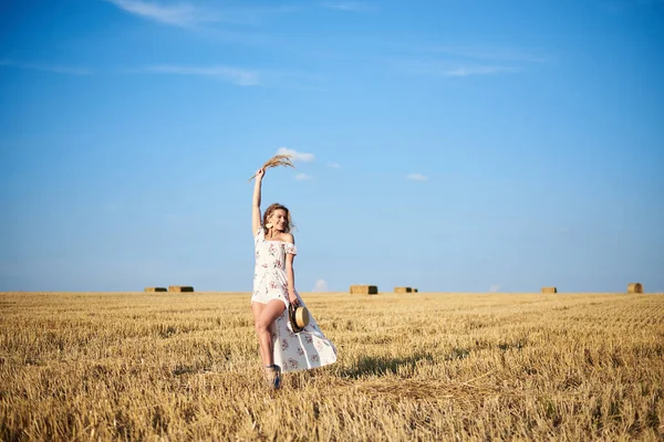 Genç sarışın kadın, beyaz romantik elbise ve hasır şapka giyiyor, elinde kuru çim buketi tutuyor, yazın saman tarlasında yürüyor. Doğal arka planda bir kadın portresi. Çevre koruma kavramı.