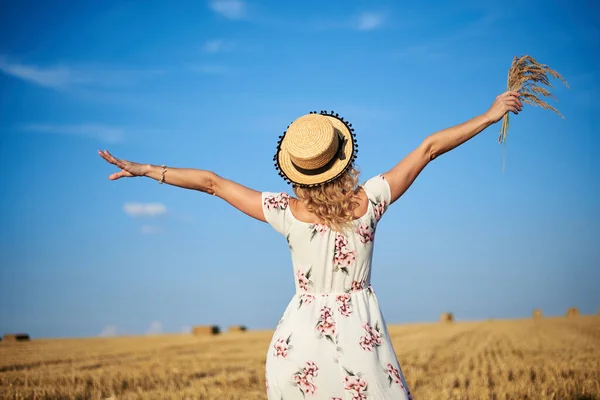年轻的金发女人 穿着白色的浪漫衣服 头戴草帽 背对着相机 两臂叉腰 夏天走在草场上 自然背景的女性肖像 — 图库照片