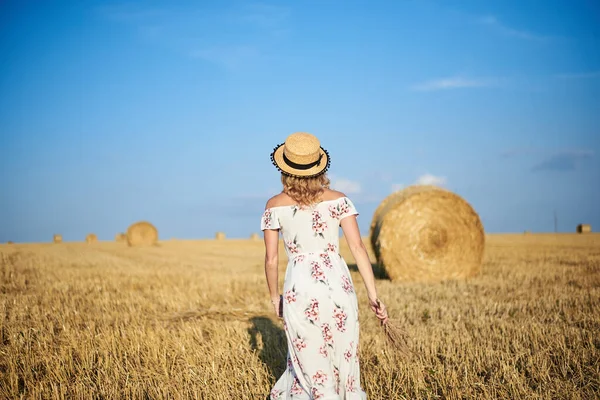 Genç sarışın kadın, beyaz romantik elbise ve hasır şapka ve kurutulmuş çim buketi giyiyor, yazın saman tarlasında koşuyor. Doğal arka planda bir kadın portresi. Çevre koruma.