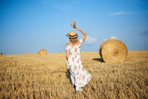 Genç sarışın kadın, beyaz romantik elbise ve hasır şapka ve kurutulmuş çim buketi giyiyor, yazın saman tarlasında koşuyor. Doğal arka planda bir kadın portresi. Çevre koruma.