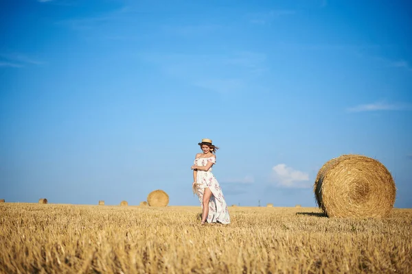 Genç sarışın kadın, beyaz romantik elbise ve hasır şapka ve kurutulmuş çim buketi giyiyor, yazın saman tarlasında koşuyor. Doğal arka planda bir kadın portresi. Çevre koruma.