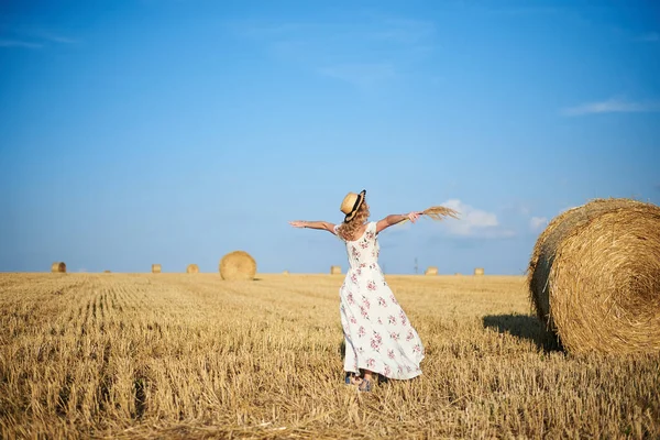 Genç sarışın kadın, beyaz romantik elbise ve hasır şapka ve kurutulmuş çim buketi giyiyor, yazın saman tarlasında koşuyor. Doğal arka planda bir kadın portresi. Çevre koruma.