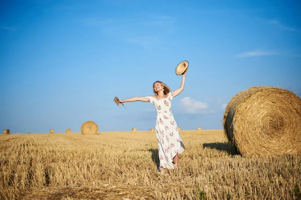Beyaz romantik elbiseler giyen, elinde hasır şapka ve kurutulmuş çim buketi olan genç sarışın kadın yazın saman tarlasında zıplıyor. Doğal arka planda bir kadın portresi. Çevre koruma.