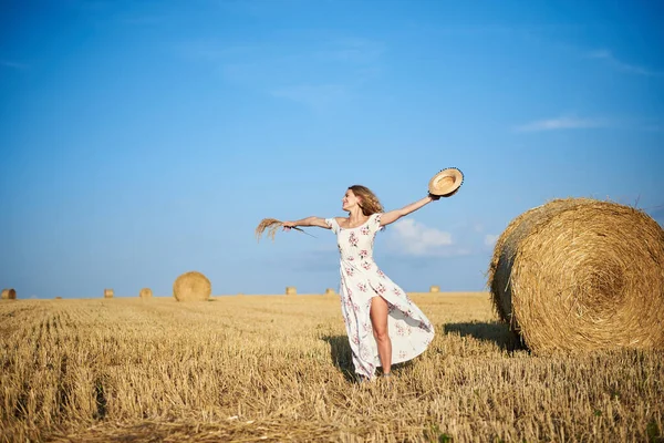 Beyaz romantik elbiseler giyen, elinde hasır şapka ve kurutulmuş çim buketi olan genç sarışın kadın yazın saman tarlasında zıplıyor. Doğal arka planda bir kadın portresi. Çevre koruma.