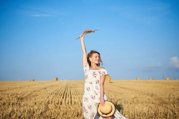 Beyaz romantik elbiseler giyen, elinde hasır şapka ve kurutulmuş çim buketi olan genç sarışın kadın yazın saman tarlasında zıplıyor. Doğal arka planda bir kadın portresi. Çevre koruma.