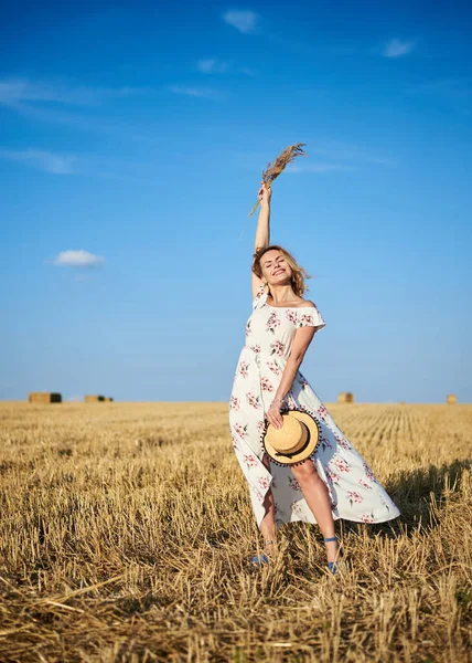 Beyaz romantik elbiseler giyen, elinde hasır şapka ve kurutulmuş çim buketi olan genç sarışın kadın yazın saman tarlasında zıplıyor. Doğal arka planda bir kadın portresi. Çevre koruma.