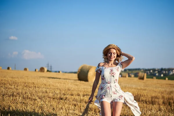 Genç sarışın kadın, beyaz romantik elbise ve hasır şapka ve kurutulmuş çim buketi giyiyor, yazın saman tarlasında koşuyor. Doğal arka planda bir kadın portresi. Çevre koruma.