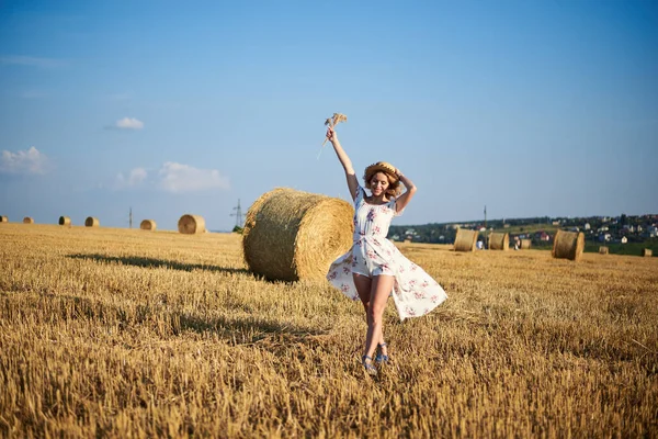 Genç sarışın kadın, beyaz romantik elbise ve hasır şapka ve kurutulmuş çim buketi giyiyor, yazın saman tarlasında koşuyor. Doğal arka planda bir kadın portresi. Çevre koruma.