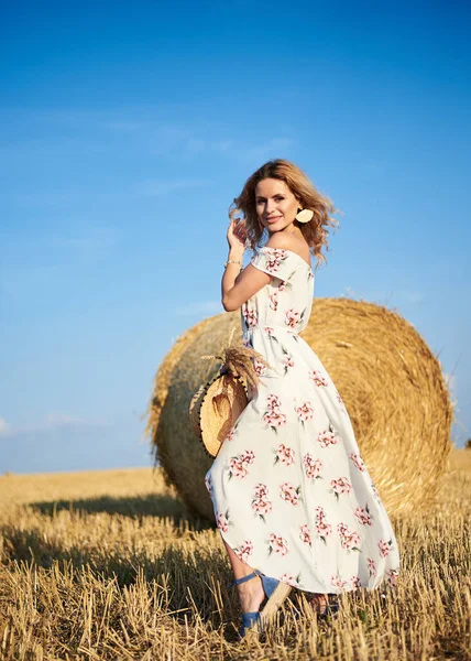 Sarışın, uzun, romantik bir elbise giyen, elinde hasır şapka ve kurutulmuş çimen buketi tutan, yazın tarlada buğday balyasının önünde poz veren genç bir kadın. Doğal arka planda kadın portresi.