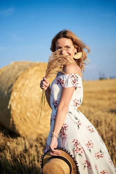 年轻的金发女人 穿着长长的白色浪漫连衣裙 头戴草帽 头戴干草束 夏天站在田边麦包前 自然背景的女性肖像 — 图库照片