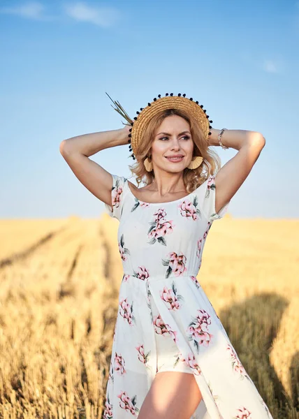 Uzun, romantik bir elbise ve hasır şapka giyen, elinde kurutulmuş çimen buketi tutan, yazın sarı alanda poz veren genç sarışın bir kadın. Doğal arka planda kadın portresi.