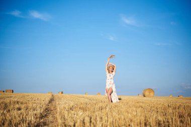 Genç sarışın kadın, beyaz romantik elbise ve hasır şapka ve kurutulmuş çim buketi giyiyor, yazın saman tarlasında yürüyor. Doğal arka planda bir kadın portresi. Çevre koruma.