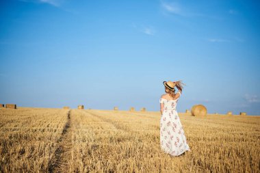 Genç sarışın kadın, beyaz romantik elbise ve hasır şapka ve kurutulmuş çim buketi giyiyor, yazın saman tarlasında yürüyor. Doğal arka planda bir kadın portresi. Çevre koruma.