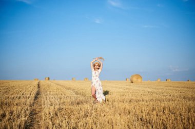 Genç sarışın kadın, beyaz romantik elbise ve hasır şapka ve kurutulmuş çim buketi giyiyor, yazın saman tarlasında yürüyor. Doğal arka planda bir kadın portresi. Çevre koruma.