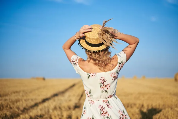 若いブロンドの女性は 白いロマンチックなドレスを着て バックカメラには 藁帽子を保持し 頭の上に草の花束を乾燥させ 夏にはわら畑を歩く 自然を背景にした女性の肖像 — ストック写真