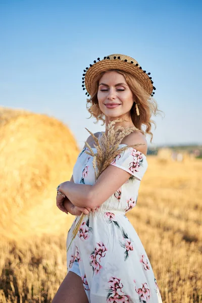 Sarışın, uzun, romantik bir elbise ve hasır şapka giyen, elinde kurutulmuş çim buketi tutan yazın tarlada buğday balyasının önünde poz veren genç bir kadın. Doğal arka planda kadın portresi.