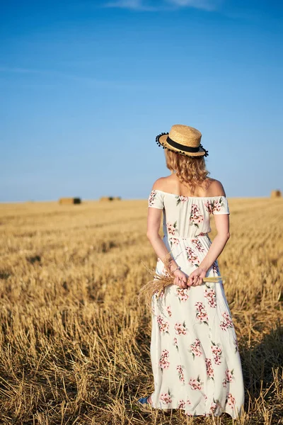 年轻的金发女人 身穿白色的浪漫衣服 头戴草帽 背对着相机 夏天时 她拿着干草束站在草场上 自然背景的女性肖像 — 图库照片