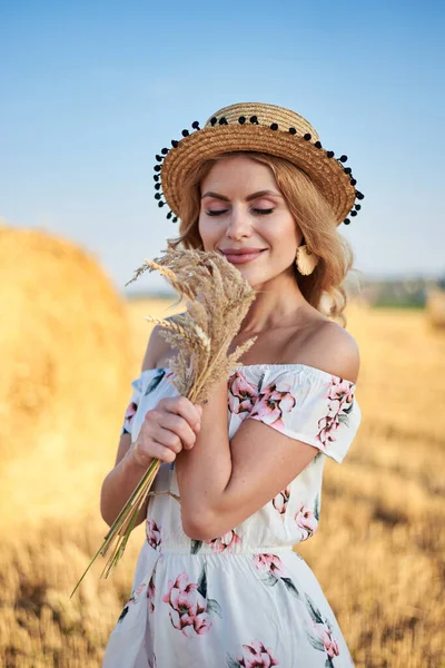 Sarışın, uzun, romantik bir elbise ve hasır şapka giyen, elinde kurutulmuş çim buketi tutan yazın tarlada buğday balyasının önünde poz veren genç bir kadın. Doğal arka planda kadın portresi.
