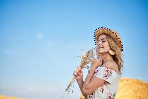 若いブロンドの女性は 長い白いロマンチックなドレスとわらの帽子をかぶって 夏にフィールド上の小麦の玉の前でポーズをとって 乾燥した草の花束を保持しています 自然を背景にした女性の肖像 — ストック写真