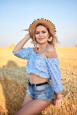 Genç kıvırcık sarışın kadın, kot şort giyiyor, açık mavi gömlek ve hasır şapka giyiyor, yazın sahada poz veriyor. Doğal kırsal kesimde bir kadın portresi. Çevre eko turizmi kavramı.