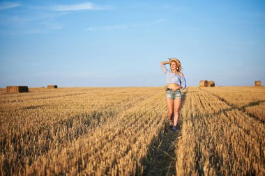 Genç kıvırcık sarışın kadın, kot şort giyiyor, açık mavi gömlek ve hasır şapka giyiyor, yazın sahada koşuyor. Doğal kırsal kesimde bir kadın portresi. Çevre eko turizmi kavramı.