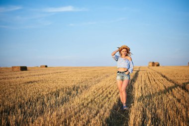 Genç kıvırcık sarışın kadın, kot şort giyiyor, açık mavi gömlek ve hasır şapka giyiyor, yazın sahada koşuyor. Doğal kırsal kesimde bir kadın portresi. Çevre eko turizmi kavramı.