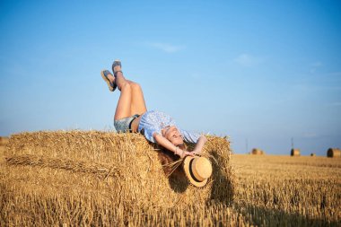 Genç kıvırcık sarışın kadın, kot şort ve açık mavi gömlek giyiyor, hasır şapka tutuyor, yazın balyada yatıyor. Doğal kırsal kesimde bir kadın portresi. Çevre eko turizmi kavramı.