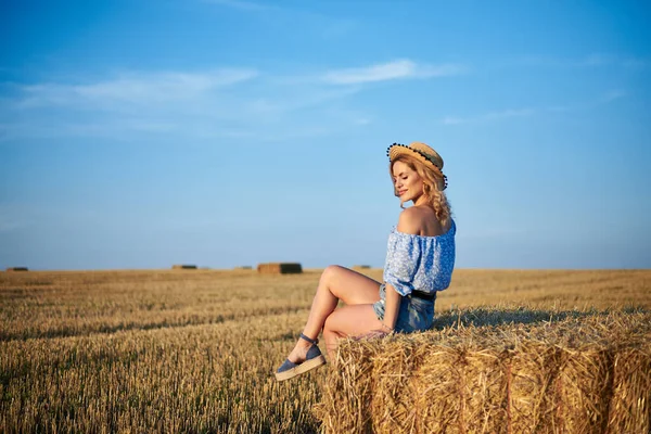 年轻的卷曲金发女人 穿着牛仔裤短裤 浅蓝色的衬衫和草帽 夏天坐在草地上的小包上 自然乡村场景中的女性肖像 环境生态旅游概念 — 图库照片