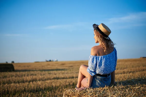 若い巻きブロンドの女性は ジーンズのショートパンツ ライトブルーのシャツとわらの帽子を着て 夏にフィールド上のカメラに戻ると座っている 自然農村の女性の肖像画 環境エコツーリズムの概念 — ストック写真