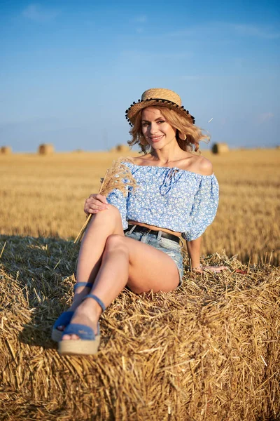 年轻的卷曲金发女人 穿着牛仔裤短裤 浅蓝色的衬衫和草帽 夏天坐在草地上的小包上 自然乡村场景中的女性肖像 环境生态旅游概念 — 图库照片