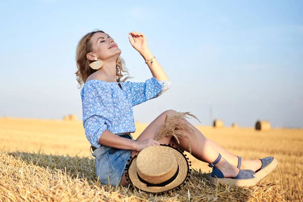 若い巻きブロンドの女性は ジーンズのショートパンツとライトブルーのシャツを着て 麦藁帽子を保持し 夏にフィールド上のベールに座っています 自然の田園風景の女性の肖像画 環境生態観光の概念 — ストック写真