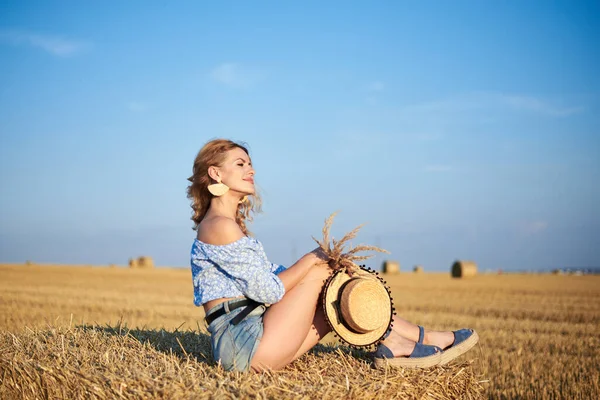 年轻的卷曲金发女人 穿着牛仔裤短裤和浅蓝色衬衫 头戴草帽 夏天坐在草地上的小包上 自然乡村场景中的女性肖像 环境生态旅游概念 — 图库照片