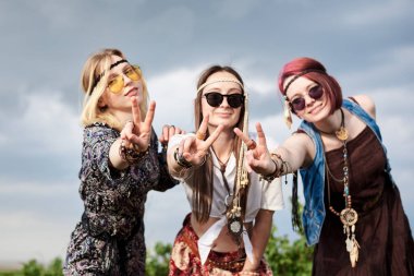 Üç hippi kadın, boho tarzı kıyafetler giyiyorlar, yeşil frenk üzümü tarlasında barış işareti gösteriyorlar, eğleniyorlar. Kırsalda birlikte seyahat eden bayan arkadaşlar. Eko turizm konsepti. Yaz tatili boş zaman