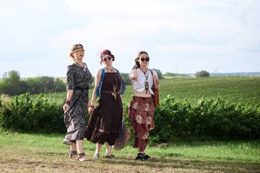 Üç hippi kadın, boho tarzı kıyafetler giyiyorlar, yeşil alanda toprak yolda yürüyorlar, eğleniyorlar. Kırsalda birlikte seyahat eden bayan arkadaşlar. Eko turizm konsepti. Yaz tatili boş zaman.