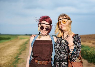 İki hippi kadın, boho tarzı kıyafetler giyiyorlar, yeşil alanda toprak yolda yürüyorlar, eğleniyorlar. Kırsalda birlikte seyahat eden bayan arkadaşlar. Eko turizm konsepti. Yaz tatili boş zaman.