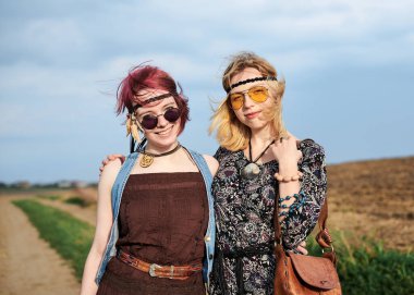 İki hippi kadın, boho tarzı kıyafetler giyiyorlar, yeşil alanda toprak yolda yürüyorlar, eğleniyorlar. Kırsalda birlikte seyahat eden bayan arkadaşlar. Eko turizm konsepti. Yaz tatili boş zaman.